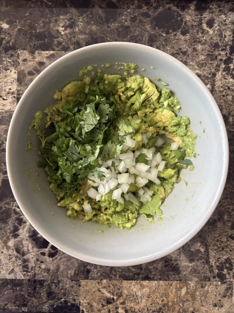 Top view of mashed avocado, finely chopped onion, and chopped cilantro leaves in a bowl.